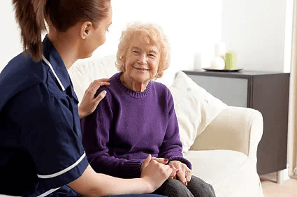 A caregiver in uniform sits with a resident on a white sofa, holding her hands in a bright, comfortable room